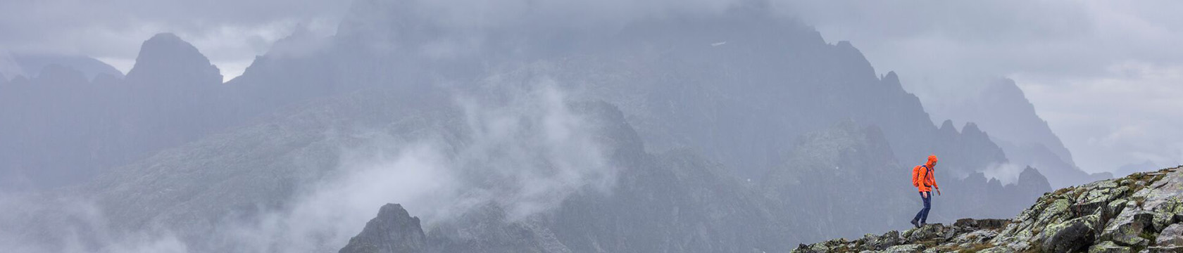 Een persoon loopt hoog in de bergen, omgeven door bergtoppen en wolken. 