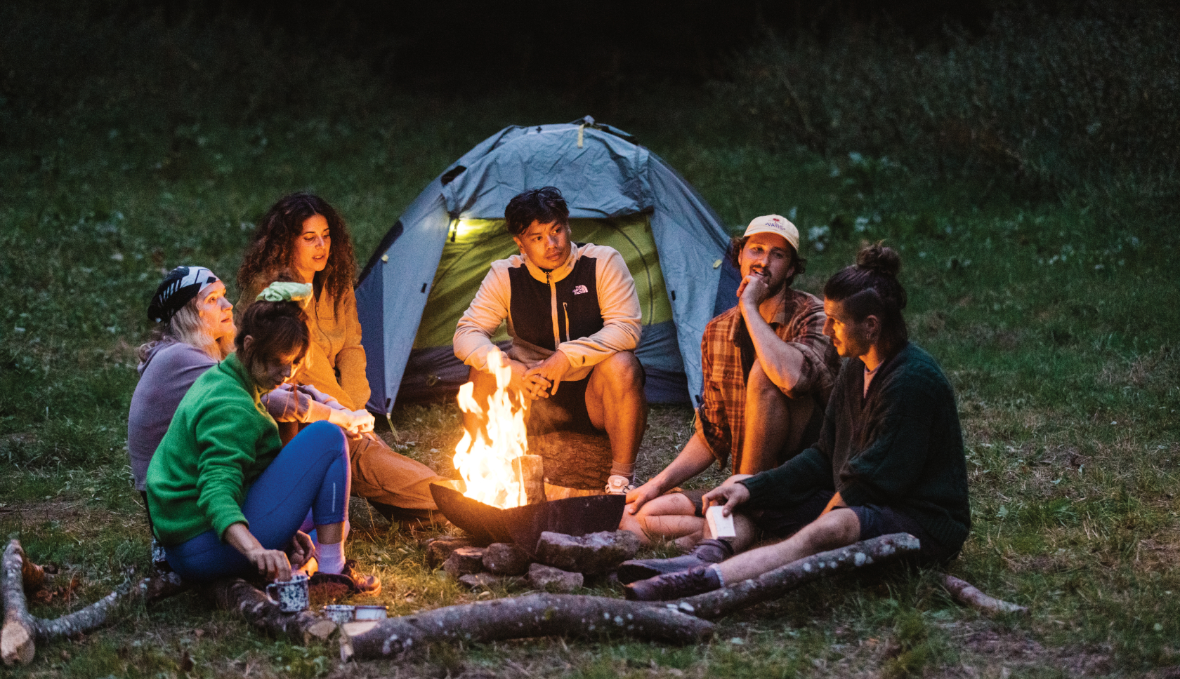 Een groep jongvolwassenen zit voor een tent rond het kampvuur