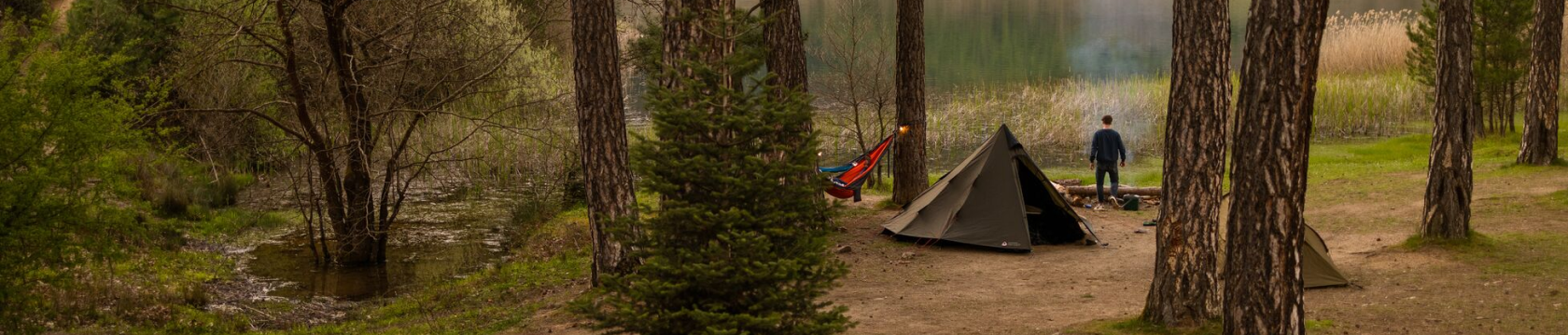 Een tipi-tent staat in het bos aan een meer, met daarnaast een man en een hangmat