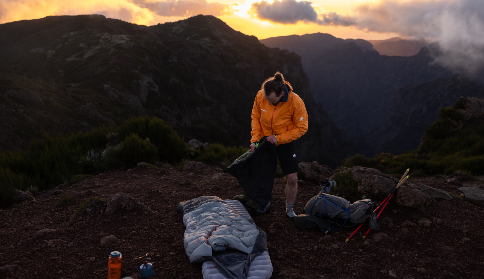 Een man is omringd door kampeerspullen bij zonsondergang op een berg