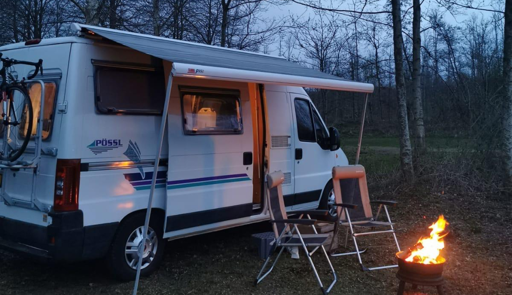 Twee stoelen staan bij een kampvuurtje voor een camperbusje