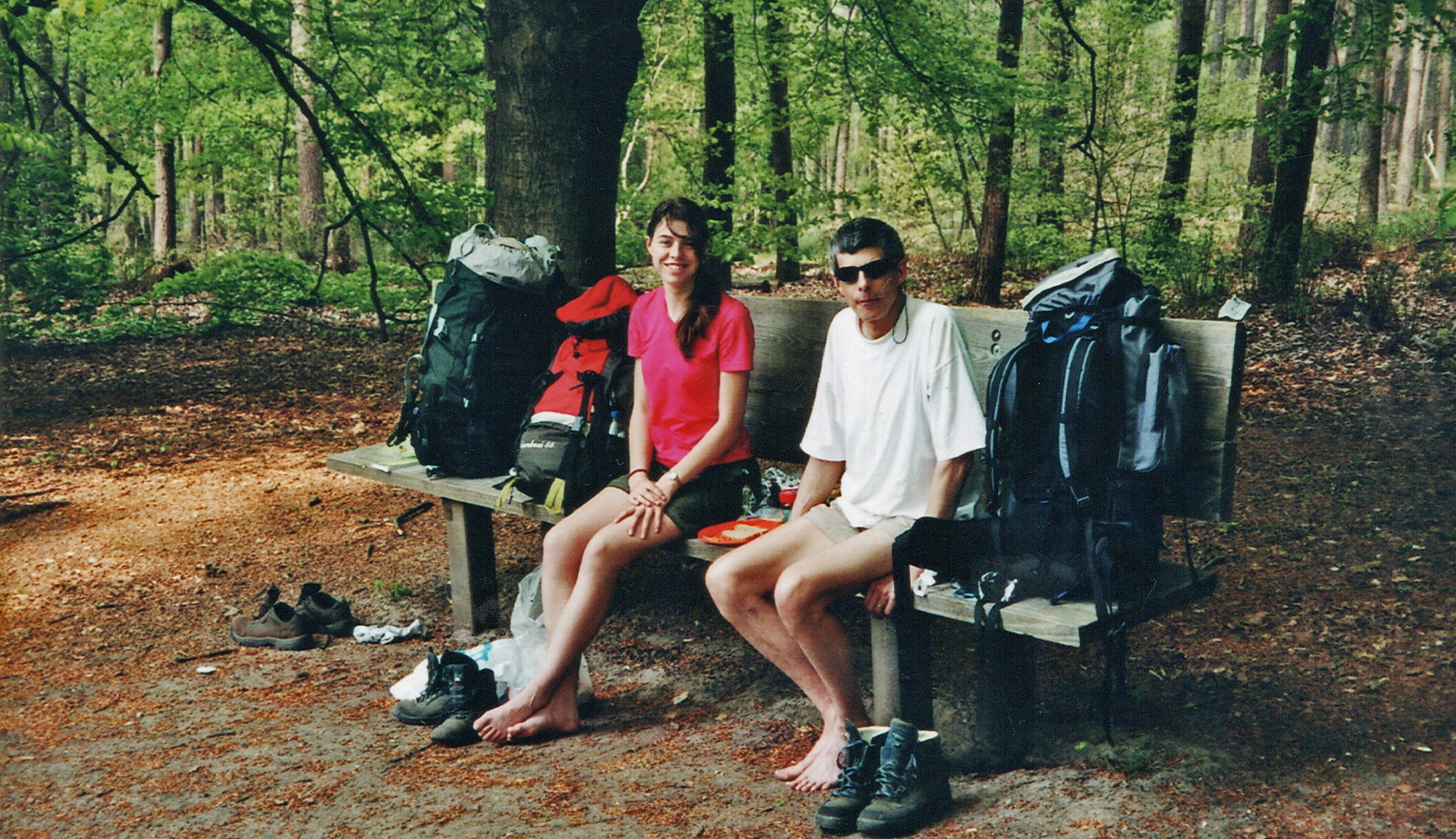 Een jonge vrouw zit blootvoets naast haar vader op een bankje, met naast hen trekkingpacks