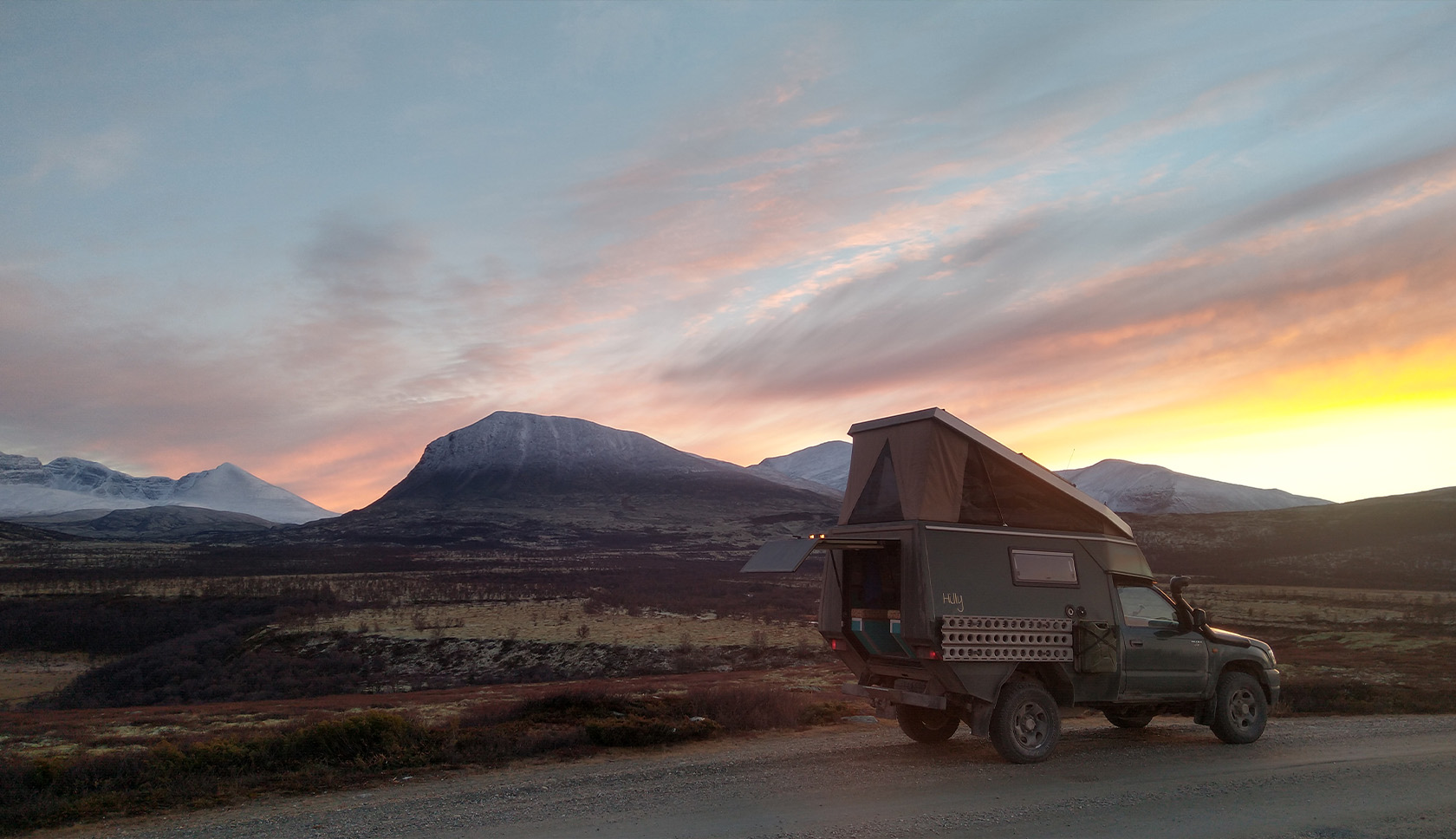 Een overlander staat geparkeerd in een heuvelachtig landschap, afgetekend tegen de felle kleuren van de ondergaande zon