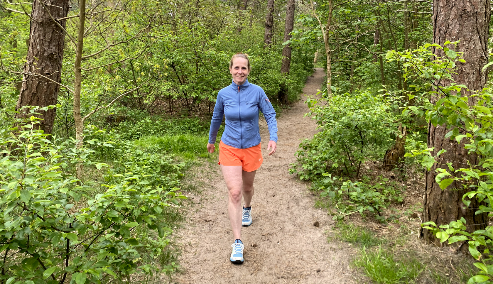 Een glimlachende vrouw in een sportieve outfit loopt in de bossen over een zandpad