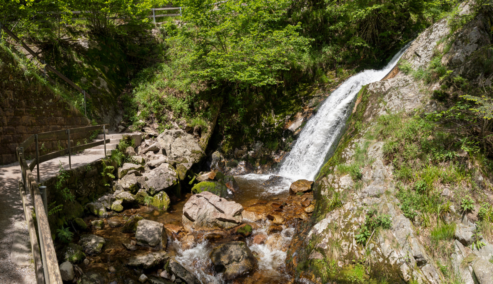 Een aangelegd pad loopt langs een waterval in een bosrijke omgeving