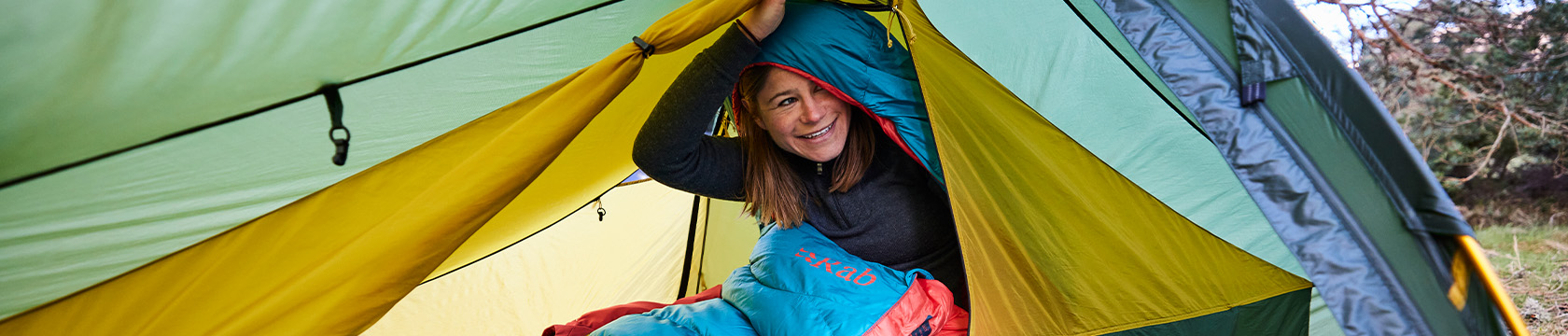 Vrouw in rab-slaapzak zit lachend in een tent